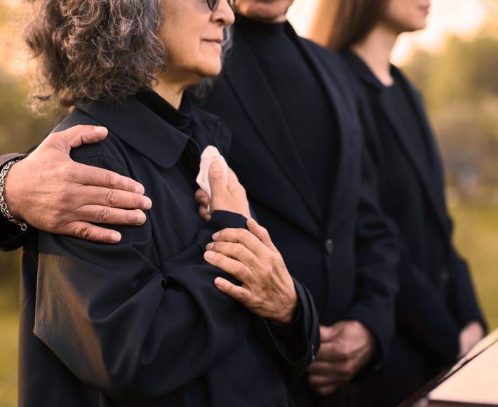 Hand of mature man on shoulder of his wife or sister with handkerchief lamenting passes away relative or family member during funeral service